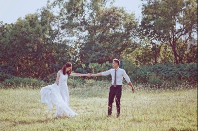 一對穿著婚紗與正式西裝的男女在草地上輕快行走，兩人手牽著手，背景為茂密的綠色樹林。