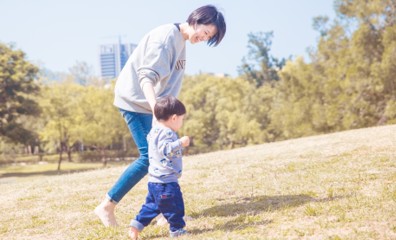 一位短髮母親在陽光明媚的公園草地上，面帶微笑地牽著年幼的孩子練習走路。